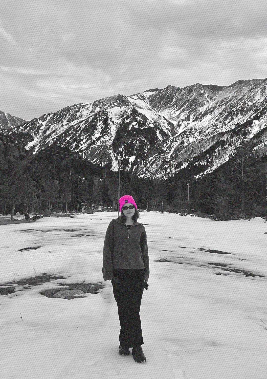 Uma fotografia em preto e branco mostra uma mulher vestindo um gorro rosa e óculos escuros, posicionada em uma paisagem nevada com montanhas ao fundo. Ela está vestida com roupas de inverno e tem uma postura relaxada.